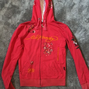Röd Ed Hardy hoodiejacka med tryck - Röd hoodiejacka från Ed Hardy med ikoniska tryck och stenar, bland annat dödskalle, rosor och blommor.  Snygga detaljer och färgstarka motiv gör den till ett statement-plagg. Storleken är M men skulle säga att den passar även S, den är använd men i väldigt bra skick, inga defekter förutom att snörena på huvan är lite uppflätade.
