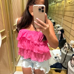 Rosa volangig linnetopp - Supersöt rosa linnetopp med flera lager volanger och tunna axelband. Toppen är croppad och har en luftig känsla, perfekt för varma dagar. Den starka rosa färgen gör att du sticker ut och volangerna ger en lekfull vibe.