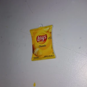 Lay's chips-påse halsbandshänge - Roligt halsbandshänge som ser ut som en Lay's Classic chips-påse. Perfekt accessoar för dig som älskar snacks och vill sticka ut. Hängsmycket är i plast och har en gul färg med Lay's logga och chipsmotiv.