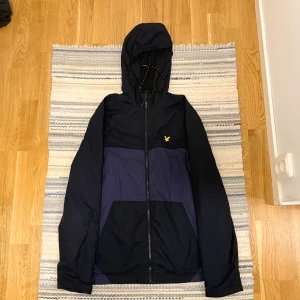 Svart och marinblå vindjacka Lyle & Scott - Snygg vindjacka från Lyle & Scott i svart och marinblått med huva och dragsko med röda detaljer. Jackan har dragkedja framtill, meshfoder och den klassiska gula loggan på bröstet. Perfekt för blåsiga dagar och streetwear-stil.