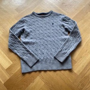 Grå kabelstickad tröja Morris M - Kabelstickad grå tröja från Morris i 100% merinoull. Klassisk rund halsringning, diskret broderad logga på bröstet och ribbade muddar. Perfekt för dig som gillar stilrena och tidlösa plagg med en modern touch. Väldigt liten i storleken och passar S