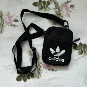 Svart axelväska från Adidas - Säljer en svart axelväska från Adidas med klassiska vita loggan framtill. Väskan har justerbar rem och dragkedja upptill samt ett extra fack med dragkedja på framsidan. Perfekt storlek för mobil, plånbok och nycklar.