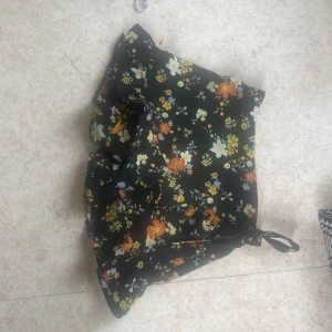 Blommiga shorts från Bik Bok XS - Supersöta svarta shorts från Bik Bok i storlek XS med färgglatt blommönster i gult, orange, vitt och lila. Modellen har volangkant och knytband i sidan för extra detalj. Perfekta för sommarens alla dagar!