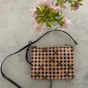 Rutig rosa axelväska med mönster - Säljer en liten axelväska med rutigt mönster i rosa, svart och brunt. Väskan har justerbar svart axelrem och stängs med dragkedja upptill. Materialet är troligtvis syntet och väskan har en stilren, fyrkantig form som passar till många outfits.