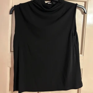 Svart polotopp från Bik Bok - En stilren svart polotopp från Bik Bok i slät polyester. Toppen är ärmlös med hög krage och har en enkel, clean look som passar till många outfits. Perfekt för dig som gillar minimalistisk stil och vill ha något som funkar året runt.