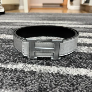 H bälte  - Snyggt vändbart bälte i skinn med klassiskt H-spänne i silver. Ena sidan är vit och den andra svart, vilket gör det enkelt att matcha med olika outfits. Bältet har en bred, stilren design och exklusiv känsla.