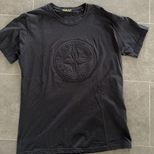 Svart t-shirt från Stone Island - Svart t-shirt från Stone Island med stort broderat logomärke på bröstet. Klassisk rund hals och korta ärmar. Skön passform och stilren design som passar perfekt till jeans eller shorts.