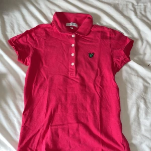 Rosa pikétopp från Lyle & Scott - Säljer en snygg rosa pikétopp från Lyle & Scott med klassisk krage, korta ärmar och knappar framtill. Toppen har en broderad grön logga på bröstet och är gjord i mjuk bomull. Perfekt för en sportig och stilren look. Andvänd runt 2-3 gånger .