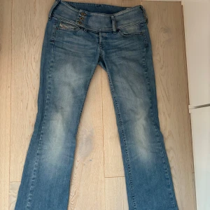 Ljusblå bootcut jeans från Diesel - Säljer ett par ljusblå bootcut jeans från Diesel med snygga broderade detaljer i guld på bakfickorna. Jeansen har låg midja, tre knappar framtill och klassisk femficksdesign. Perfekta för dig som gillar en avslappnad men ändå trendig look. Midjemåtten rakt över är 37 cm och innerbenslängden är 79cm.☺️