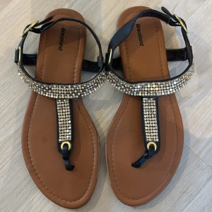 Svarta sandaler med strass från Graceland - Snygga svarta sandaler från Graceland med bruna sulor och glittrande strassdetaljer över foten. Remmarna har guldfärgade spännen och sandalerna har platt sula. .