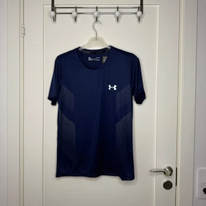 Blå Under Armour tränings-tshirt M - Snygg marinblå t-shirt från Under Armour med vit logga på bröstet och tryckt text på ryggen. Gjord i ett lätt och snabbtorkande polyestermaterial, perfekt för träning. T-shirten har korta ärmar och diskreta mönsterdetaljer på sidorna.