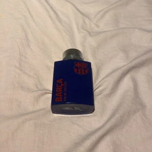 Barça Eau de Toilette 100ml - Parfymflaska i mörkblått glas med FC Barcelonas logga och text i rött. Flaskan har en robust, silvrig metallkork med räfflad yta. Innehåller 100 ml Eau de Toilette, perfekt för dig som älskar fotboll och vill ha en sportig doft. Den finns inte och köpa längre.