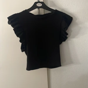 Svart volangblus från Zara - Supersnygg svart blus från Zara med volangdetaljer på axlarna. Modellen är croppad och har en stilren, enkel design som passar perfekt till jeans eller kjol. Materialet känns mjukt och följsamt mot huden.