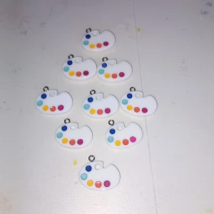 Färgglada palettberlocker i plast - Säljer små vita berlocker formade som konstnärspaletter med färgglada prickar i blått, rött, orange, gult och rosa. Perfekta att använda till halsband, armband eller örhängen för en kreativ och lekfull stil.