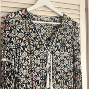 Blommig kortklänning  - Supersöt kortklänning med blommigt mönster i marinblått, vitt och röda detaljer. Klänningen har v-ringning med snörning och långa ärmar. Storlek M 