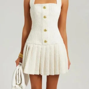 Vit kortklänning med guldknappar - Supersöt vit kortklänning i strukturerat tyg med breda axelband, markerad midja och plisserad kjol. Framsidan har stora guldknappar och baksidan en snygg guldfärgad dragkedja. Perfekt för dig som gillar en chic och trendig look.