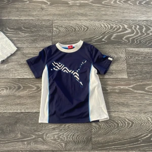 Blå och vit Puma t-shirt med zebra - Snygg t-shirt från Puma i marinblått och vitt med coolt zebramönstrat Puma-tryck på bröstet. T-shirten har korta ärmar, rund halsringning med vit ribbad kant och blå detaljer längs sidorna. Perfekt för sport eller chill vardagsstil.
