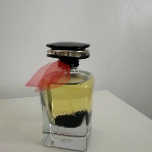 Bella Rouge Eau de Parfum - Elegant parfymflaska med en svart och silverfärgad kork. Flaskan är dekorerad med ett rött band runt halsen och innehåller en ljusgul vätska. Perfekt för den som söker en sofistikerad doftupplevelse.