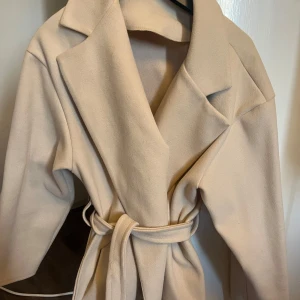 Beige kappa med bälte - Elegant beige kappa med bälte i midjan. Den har en klassisk krage och långa ärmar, perfekt för vår/höst eller kallare sommarkvällar från Missäy i storlek s/m. 