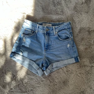 Blå jeansshorts från Bershka - Snygga blå jeansshorts från Bershka med slitna detaljer och uppvikta kanter. Perfekta för sommardagar och ger en avslappnad look. De har fem fickor och en knappgylf.