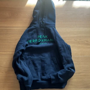 Mörkblå hoodie från Peak Performance - Säljer en snygg mörkblå hoodie från Peak Performance med grön text på bröstet. Den har en klassisk känguruficka och en bekväm huva. Perfekt för kyliga dagar!