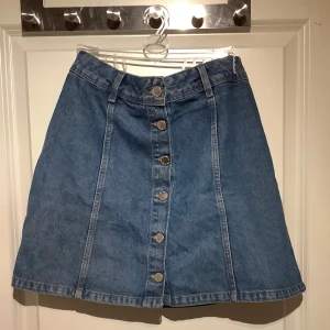 Jeanskjol från H&M Divided - Snygg jeanskjol från H&M Divided med knappar framtill. Kjolen har en klassisk blå denimfärg och en A-linjeformad siluett. Perfekt för en avslappnad stil.