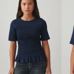 Slutsåld mörkblå topp från Gina Tricot - Säljer en mörkblå topp från Gina Tricot med korta ärmar och en smockad design framtill. Toppen har en söt volangdetalj vid nederkanten. Perfekt för en stilren look! Den är endast använd en gång. Pris går att diskutera. 