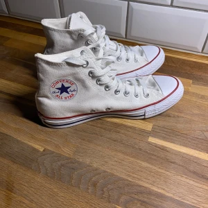 Vita höga sneakers från Converse - Säljer ett par klassiska vita höga sneakers från Converse med snörning och rund tå. Skorna har den ikoniska loggan på sidan och en röd rand längs sulan. Perfekta för en avslappnad stil.