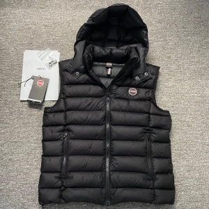 Svart dunväst från Colmar - Snygg svart dunväst från Colmar med avtagbar huva och dragkedja framtill. Västen har två praktiska fickor med dragkedja och en stilren design. Perfekt nu till våren! Finns en liten lagning (bild 3) annars är den i perfekt skick🍾Nypris ca 3500 