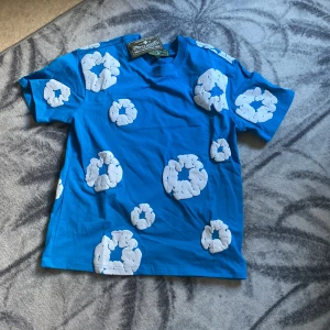 Blå t-shirt med vita mönster från Denim Tears - Säljer en  blå t-shirt från Denim Tears med unika vita mönster över hela plagget. Perfekt för att sticka ut i mängden. Gjord i mjukt material för en bekväm känsla hela dagen.