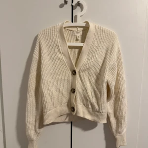 Vit stickad kofta från H&M - Säljer en stilren vit stickad kofta från H&M med stora knappar framtill. Koftan har en v-ringad design och långa ärmar, perfekt för kyligare dagar. Den är mjuk och bekväm att bära. Passar bra till både jeans och kjol.