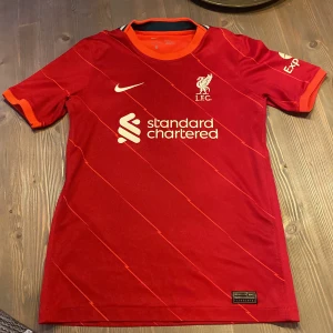 Röd liverpool tröja från Nike - Säljer en röd fotbollströja från Nike med LFC-logga och sponsortryck. Tröjan har korta ärmar och ett subtilt randigt mönster. Perfekt för fotbollsfans! 🔴