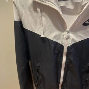 Svart och vit vindjacka från Nike - Säljer en snygg svart och vit vindjacka från Nike med dragkedja och huva. Jackan har en klassisk design med Nike-logga på bröstet och justerbara snören vid huvan. Perfekt för blåsiga dagar!