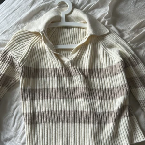 Randig ribbad tröja från NA-KD - Säljer en snygg ribbad tröja från NA-KD i krämvitt och beige. Tröjan har en klassisk krage och v-ringning, perfekt för en stilren look. Den är långärmad och har ett randigt mönster som ger en modern touch.