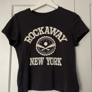 Svart t-shirt från H&M Divided - Säljer en svart t-shirt från H&M Divided med trycket 'Rockaway New York' i vitt. T-shirten har en rund hals och korta ärmar, perfekt för en casual look. Passar bra till jeans eller shorts för en avslappnad stil.