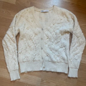 Beige / vit kofta med pärlor - Säljer en elegant beige kofta från Zara dekorerad med pärlor. Koftan har en v-ringad design och långa ärmar, perfekt för att ge en stilren touch till din outfit. Den är tillverkad i ett mjukt material som känns skönt mot huden. Perfekt för både vardag och speciella tillfällen. Har endast använt 1-2 gånger är i mycket bra skick.