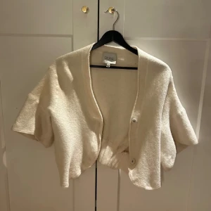 Beige kofta från Dagmar - Säljer en stilren beige kofta från Dagmar. Koftan har korta ärmar och knappar framtill. Perfekt för lager-på-lager-stil och ger en elegant touch till din outfit.