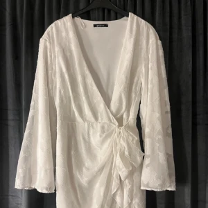 Vit omlottklänning från Gina Tricot - Elegant vit omlottklänning från Gina Tricot med v-ringning och knytning i midjan. Klänningen har ett subtilt blommönster och långa ärmar, perfekt för en stilren look. Endast använd en gång! Ny pris ca 600-700, kan diskutera priset.