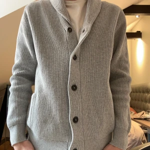 Grå ribbad kofta från M&S - Säljer en stilren grå kofta från M&S. Den är ribbad med knappar framtill och har en klassisk krage. Perfekt för sena sommarkvällar och ger en avslappnad men ändå elegant look.