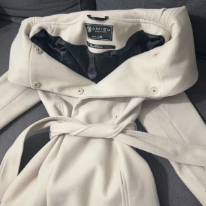 Beige kapa  - Elegant vit kappa från Amisu med knappar och bälte i midjan. Perfekt för en stilren look. Kappan har en klassisk design med långa ärmar och en mjuk krage.