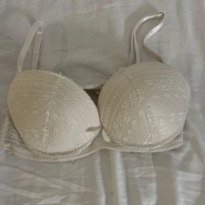 Vit spets-bh med rosett - Säljer en elegant vit bh med spetsdetaljer och en söt rosett framtill. Bh:n har justerbara axelband och en klassisk hakknäppning bak. Storlek: 75B