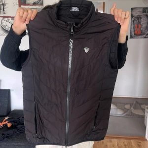 Svart quiltad väst med dragkedja - Snygg svart quiltad väst från EA7 Emporio Armani med dragkedja framtill. Perfekt för lager-på-lager under kyligare dagar. Västen har en stilren design med ett litet märke på bröstet.