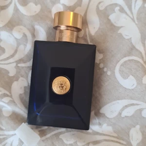 Versace Pour Homme Dylan Blue Eau de Toilette - Säljer en elegant flaska av Versace Pour Homme Dylan Blue. Flaskan är mörkblå med en gyllene kork och ett ikoniskt Versace-emblem i guld på framsidan. Perfekt för den som vill ha en sofistikerad och maskulin doft.