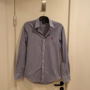 Randig skjorta från Polo Ralph Lauren - Snygg blå- och vitrandig skjorta från Polo Ralph Lauren med klassisk krage och knappar framtill. Den har långa ärmar och en broderad logotyp i rött på bröstet. Perfekt för sommaren. Nyskick. Passar S perfekt