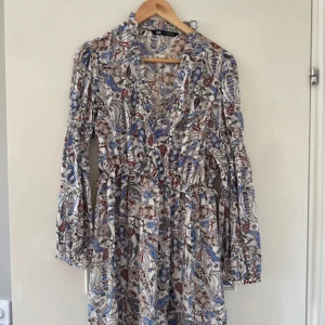 Mönstrad klänning med volangdetaljer - Säljer en vacker mönstrad klänning med volangdetaljer och v-ringning. 
