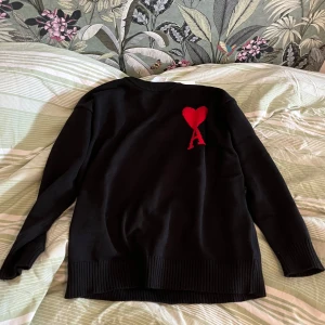 Svart tröja från Ami Paris - Säljer en svart tröja från Ami Paris med ett rött hjärta och bokstaven 'A' .Tröjan har ärmar och ribbade muddar vid ärmslut och nederkant. Perfekt för en stilren look.