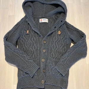 Grå stickad kofta med huva - Säljer en snygg grå stickad kofta från Viking med huva och knappar framtill. Koftan har ett flätmönster och ribbade muddar vid ärmslut och nederkant. 