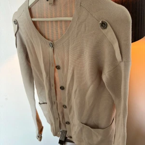 Burberry kofta  - Säljer en stilren beige kofta från Burberry med knappar framtill och dekorativa knappar på axlarna. Koftan har långa ärmar och ett klassiskt Burberry-mönster på insidan av ärmsluten. Perfekt för en elegant och avslappnad look. Självklart äkta, köptes på NK