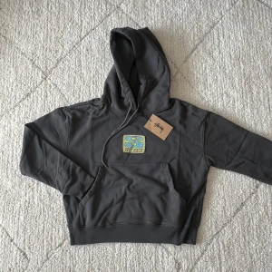 Grå hoodie från Stussy - Säljer en snygg grå hoodie från Stussy med en stor ficka framtill och justerbar huva. Den har en cool logga i blått och gult på bröstet. Perfekt för en avslappnad stil.