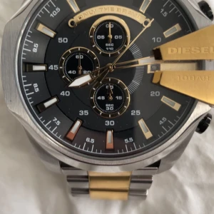 Klocka från Diesel med metallarmband - Snygg klocka från Diesel med en robust design. Den har en svart urtavla med guldiga detaljer och flera sub-dials. Armbandet är i silver och guld, vilket ger en elegant touch. Perfekt för den som gillar en stilren och modern look.